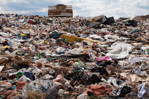 A vast, uneven landfill site filled with a mixture of accumulated waste materials, including plastic bags, paper debris, broken furniture, and miscellaneous household rubbish. The rubbish is scattered across the ground, creating a cluttered landscape with various colours, textures, and sizes. In the background, there is a partially constructed or abandoned building with a skeletal structure visible against a cloudy sky. The scene is outdoors, likely nearby a commercial or residential area, with no vegetation or natural landscape visible amidst the waste. The image emphasizes the scale and disorder of large-scale rubbish accumulation, aligning with waste management services such as those provided by Commercial Waste Whitechapel operating in the local postcode area, and it visually underscores the importance of professional rubbish removal to address such environmental issues.