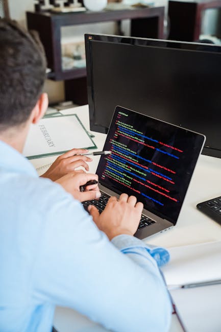 A close-up view of a person's hands typing on a laptop keyboard placed on a dark surface. The laptop screen displays lines of computer code with syntax highlighting in shades of green, blue, and white