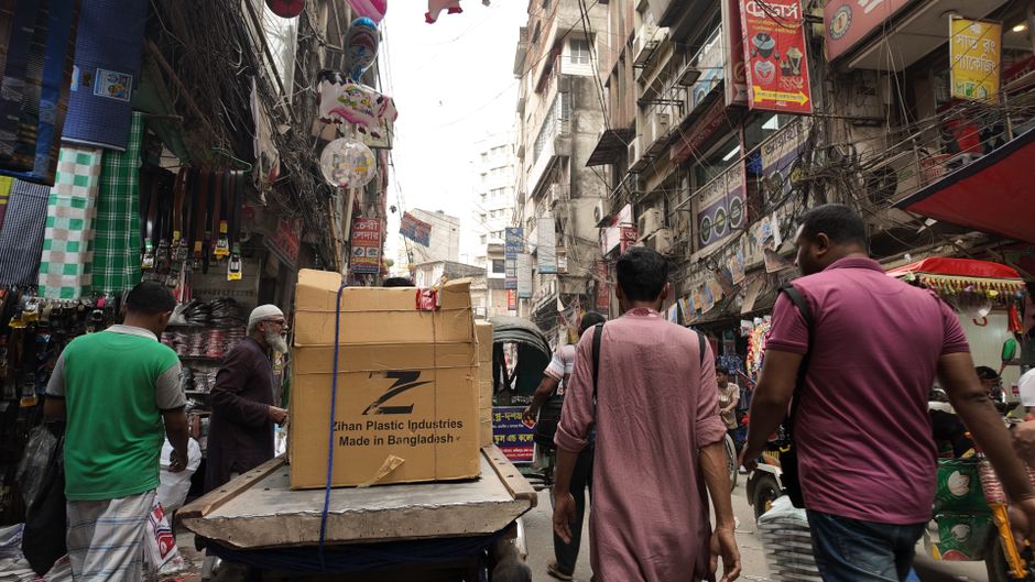 A large stack of wooden fruit boxes, primarily cardboard with printed labels reading 'fresh fruits' and colorful images of vegetables, are piled outdoors on pavement. The boxes vary in condition, with