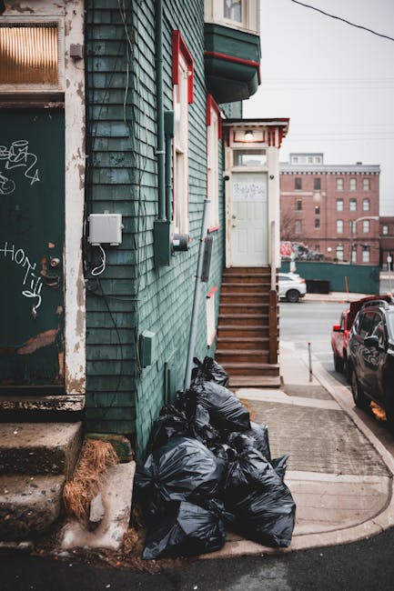 The image depicts the exterior corner of a building with weathered green wooden cladding, positioned on a street in an urban environment. Several black plastic rubbish bags are piled against the build