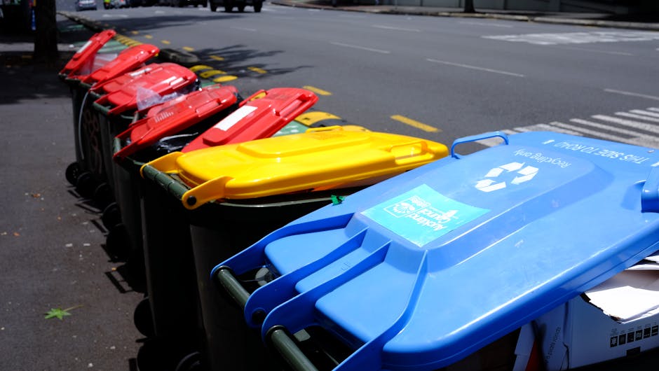 A row of waste bins arranged along the edge of a pavement on a city street, each with a different colour lid—red, yellow, and blue—indicating separate waste or recycling categories. The blue bin, 
